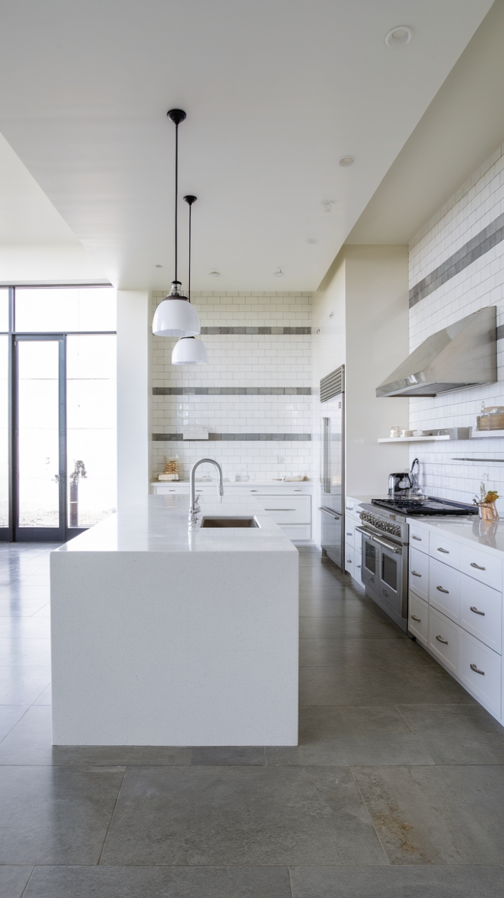 This stunning modern kitchen embodies the essence of minimalist design, where every element harmonizes to create a serene culinary haven. The expansive island, crowned with a pristine white quartz countertop, serves as both a functional workspace and a stylish gathering spot, illuminated by two elegant pendant lights that add a touch of sophistication. Surrounding the island, sleek white cabinets and a chic backsplash adorned with subtle grey accents enhance the kitchen's bright and airy atmosphere, while the large grey tiles underfoot provide a contemporary foundation. With top-of-the-line stainless steel appliances, including a refrigerator and a striking range hood, this kitchen is not just a place for cooking but a canvas for culinary creativity.