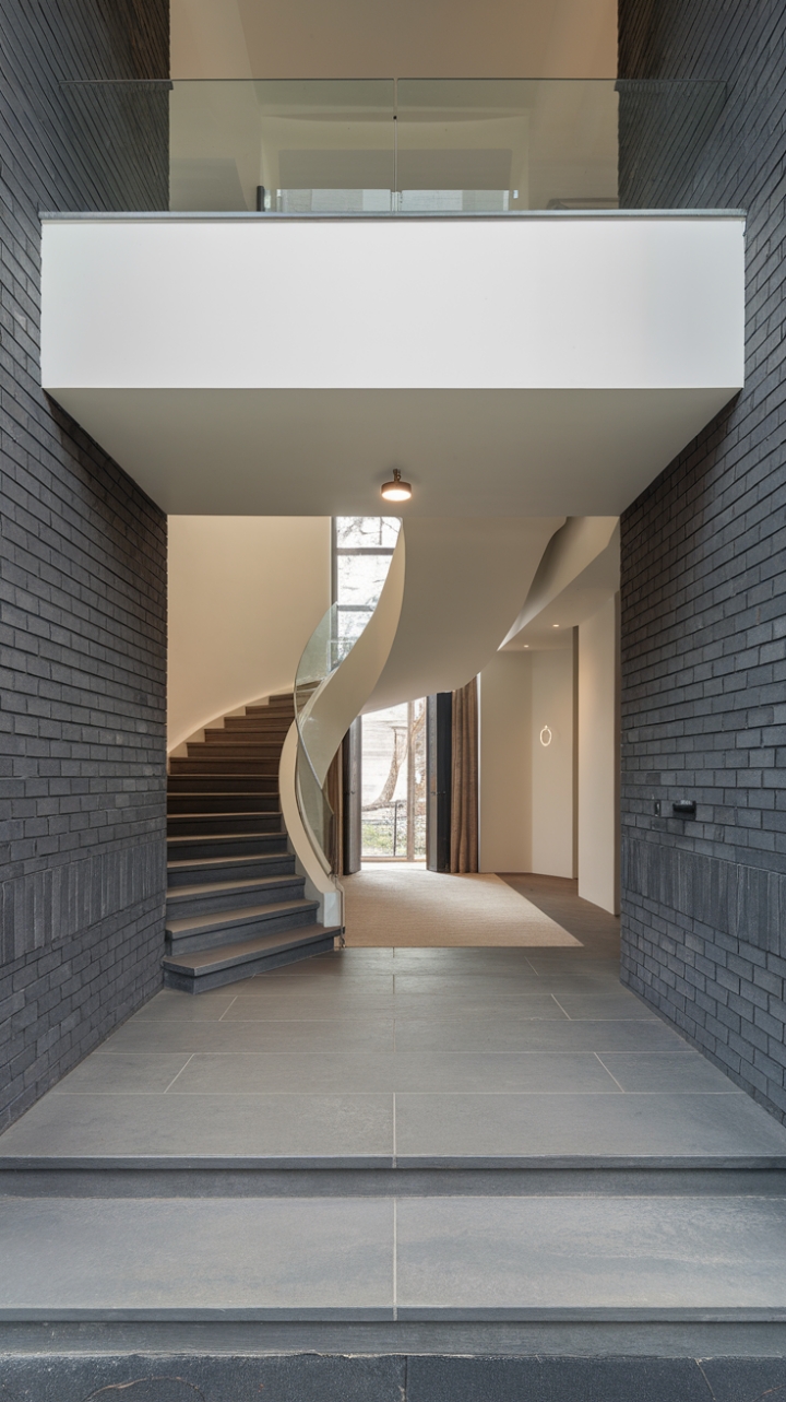A stunning blend of modern elegance and understated charm, this entrance foyer captivates with its minimalist aesthetic, featuring a gracefully curved staircase framed by a transparent glass railing that showcases the architectural beauty. The large gray tiles underfoot provide a sleek foundation, while the dark gray brick wall on the left introduces a bold contrast, its strategically placed holes adding an element of surprise. The lofty white ceiling amplifies the airy feel, with a delicate round light fixture hanging above, illuminating the path to a stylish entry corridor adorned with a plush beige carpet, inviting guests into a world of refined design.